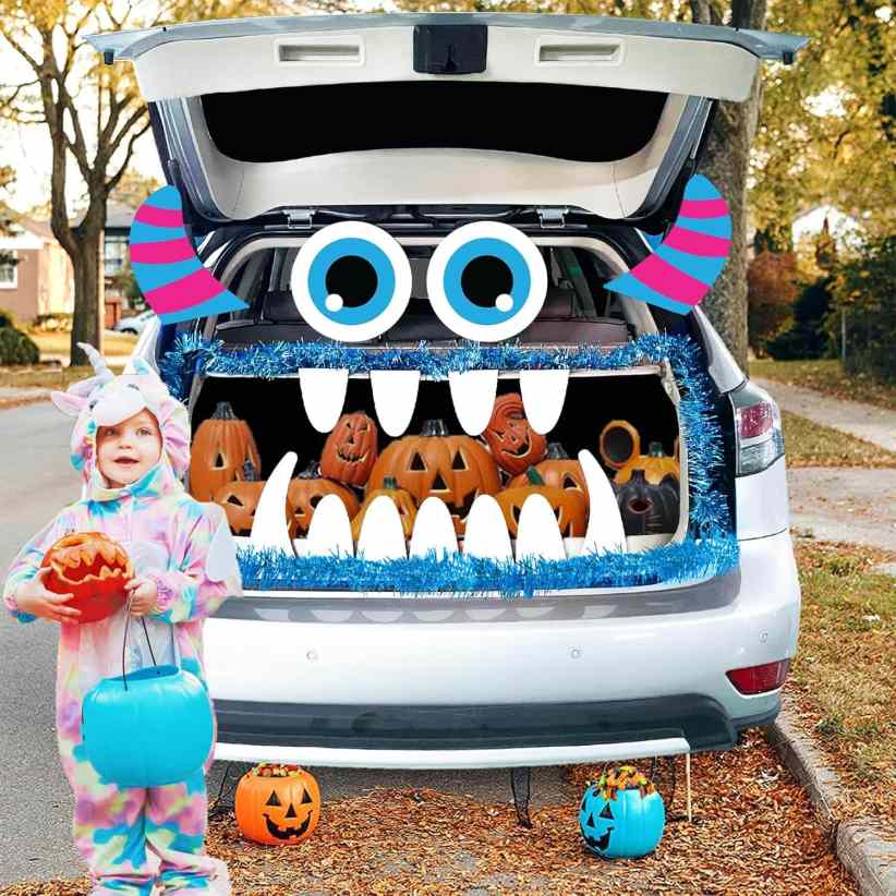 White SUV trunk decorated as a blue-eyed monster with striped horns and big teeth, filled with jack-o’-lanterns, beside a child in unicorn costume.