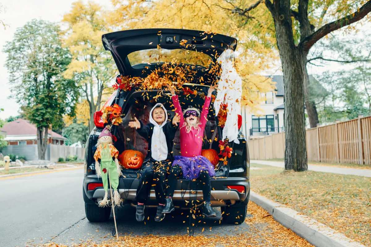 Two kids in Halloween costumes sit in an open SUV trunk decorated with pumpkins and fall leaves, joyfully tossing leaves into the air.