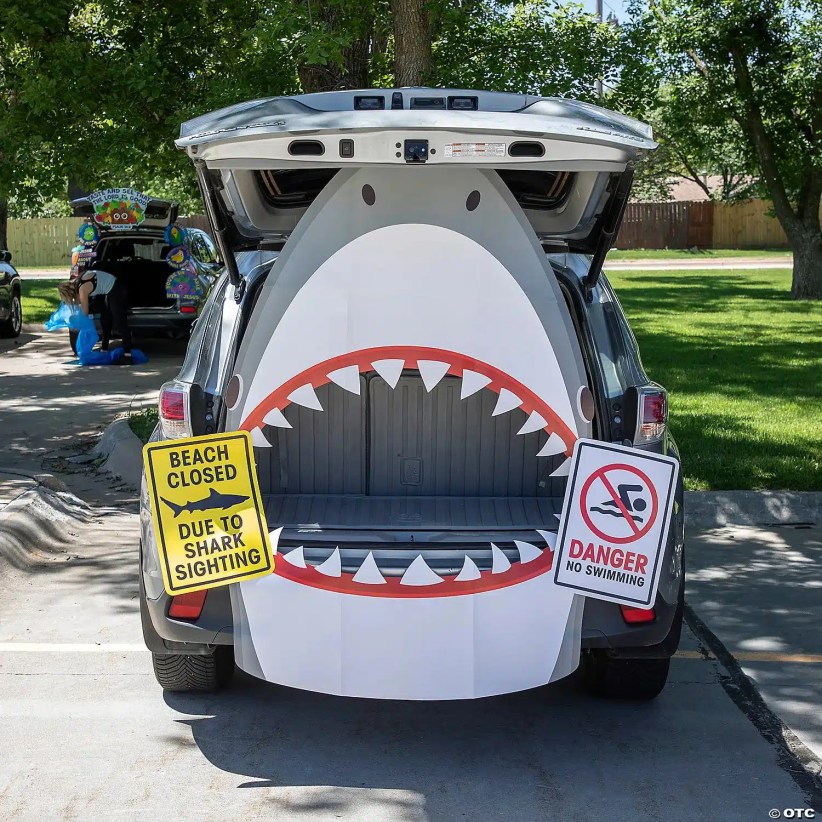 SUV trunk decorated as a gray and white shark mouth with teeth, flanked by “Beach closed” and “Danger no swimming” shark warning signs.