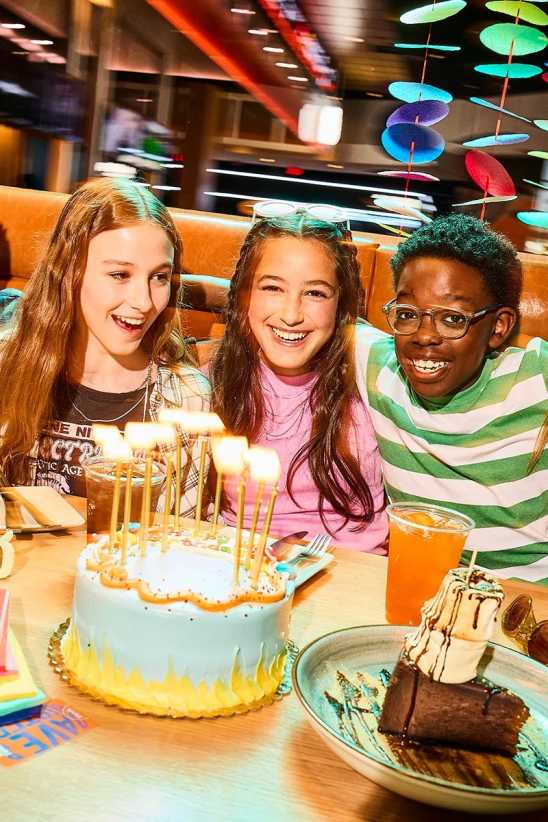 Three kids sit together in a restaurant booth celebrating a birthday, with a lit cake and desserts on the table in front of them.