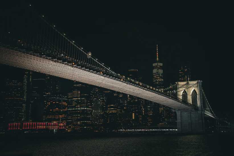 A nighttime panorama of the Brooklyn Bridge with the illuminated Manhattan skyline, including One World Trade Center.