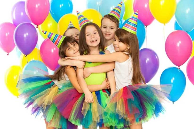Four young girls in rainbow tutus and party hats hug together, smiling in front of a colorful wall of balloons at a birthday party.