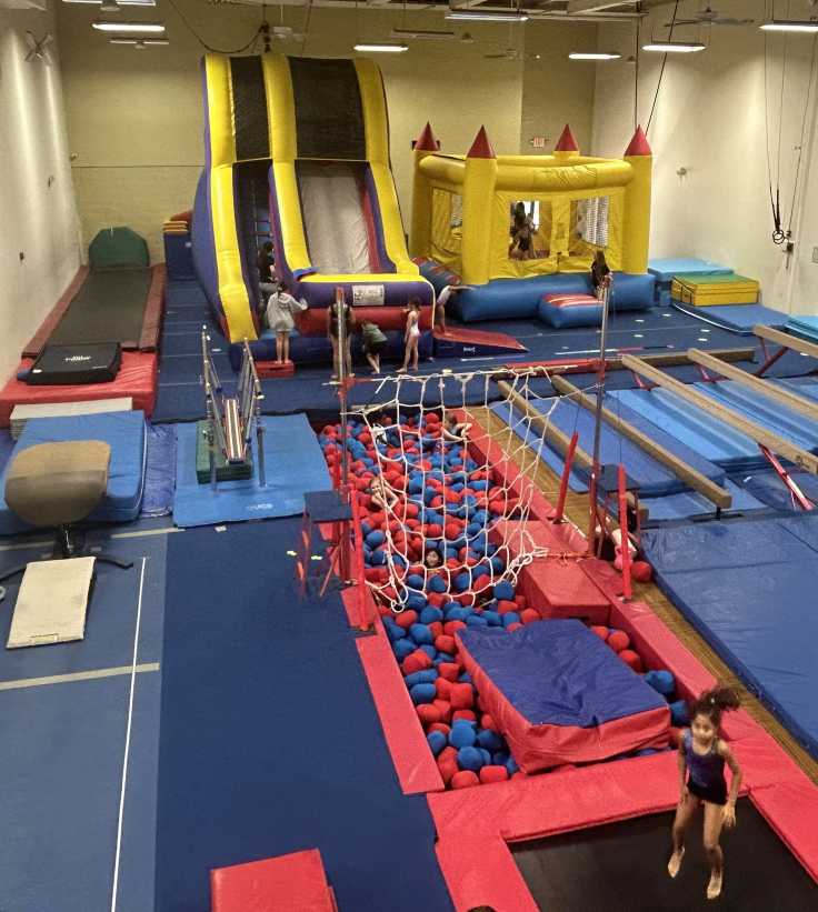 Children play in a large indoor gym filled with trampolines, balance beams, a ball pit, slides, and inflatable bounce houses.