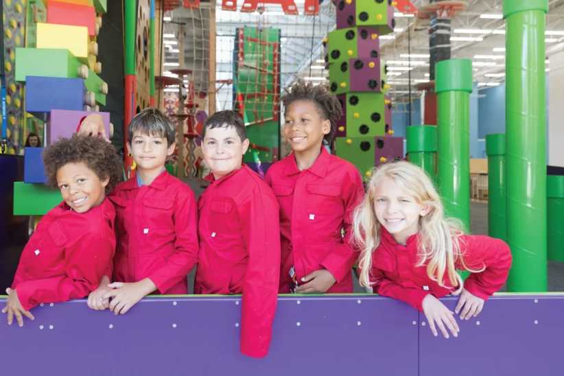 Five children in matching red jumpsuits stand and pose together at an indoor climbing gym with colorful climbing walls and play structures.