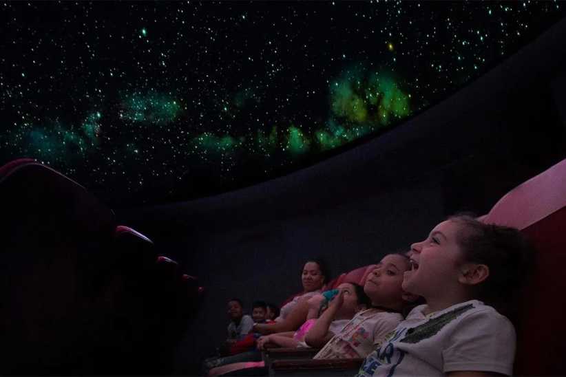 
Children lie back in reclining seats, gazing up at a domed planetarium ceiling filled with stars and glowing nebulae.