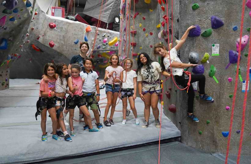
A group of children and an instructor in harnesses pose and climb at an indoor rock climbing gym, surrounded by colorful holds.