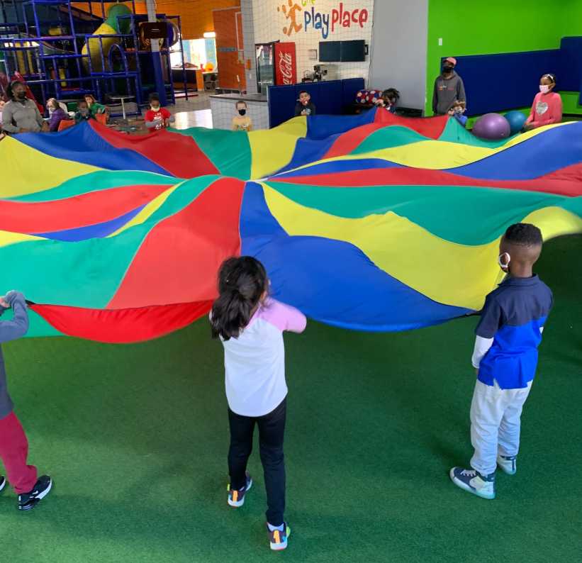 Children stand around and lift a large, colorful parachute in an indoor play center, participating in a fun group activity.
