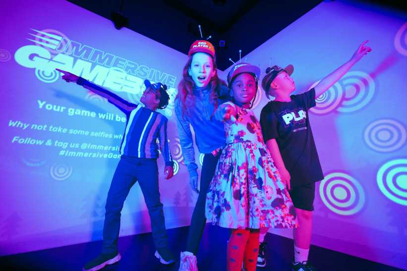 
A group of four children in colorful outfits pose and point dramatically under purple lights at an immersive gaming venue.