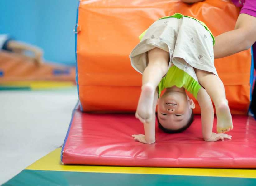 A young child in bright clothes smiles upside down while doing a handstand on a colorful gymnastics mat with an instructor nearby.