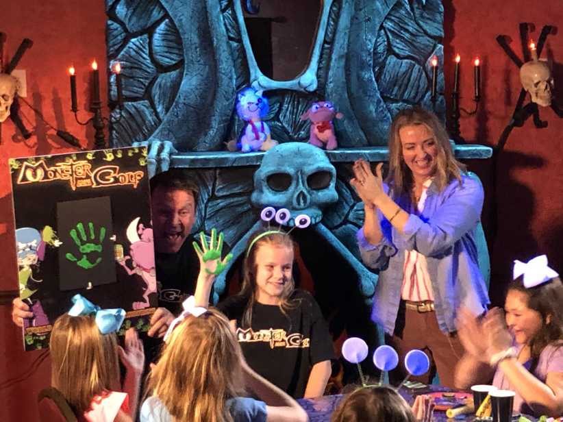 Children at a Monster Mini Golf party sit around a table with crafts; adults and decorations create a spooky, festive atmosphere.