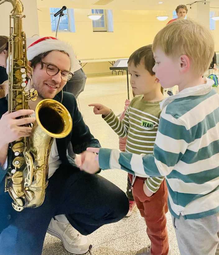 Man in Santa hat with a saxophone showing instrument to 2 toddler boys