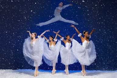 4 ballet dancers in white costumes dancing on stage while a male ballet dancer leaps over them.