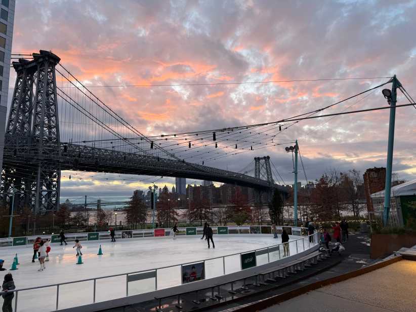 Ice skaters on a riverside rink at sunset with a large suspension bridge and colorful clouds in the background.