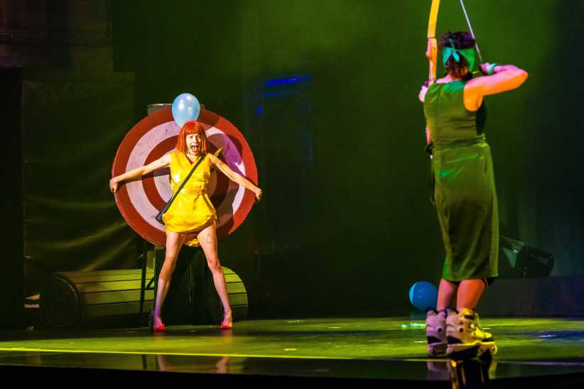 Blindfolded performer takes aim with a bow and arrow at her assistant in front of a target with a balloon over her head