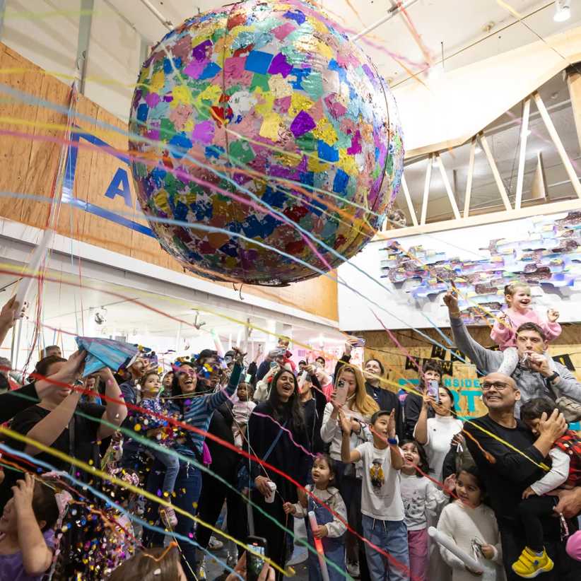 Room full of families surrounded by streamers standing under a giant ball covered in colored patches. 