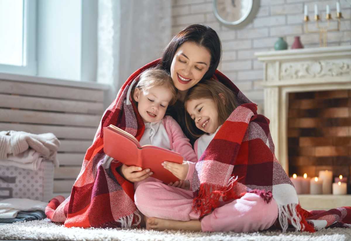 Fun portrait of happy loving family. Pretty young mother reading a winter book to her daughters at home.