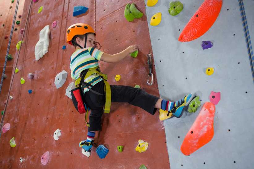 A young boy wearing a safety harness and helmet, climbing up a brightly colored wall at a rock climbing gym