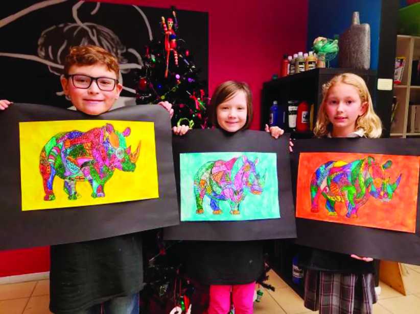 Three children proudly hold up colorful rhinoceros artwork on black mats in an art classroom, smiling at the camera.