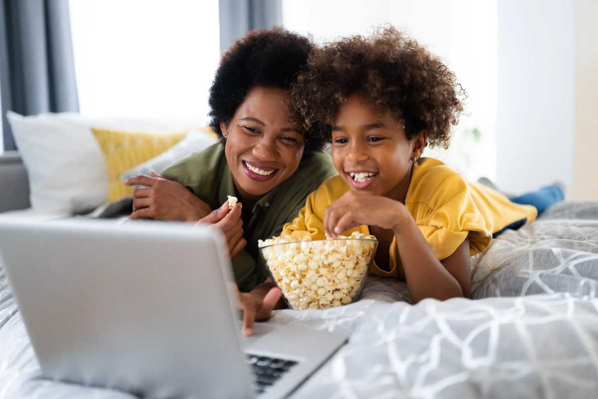 Gorgeous smiling African American single mother enjoying free time with daughter while watching funny videos online on laptop and lying on bed. Black History Month