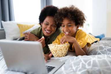 Gorgeous smiling African American single mother enjoying free time with daughter while watching funny videos online on laptop and lying on bed. Black History Month