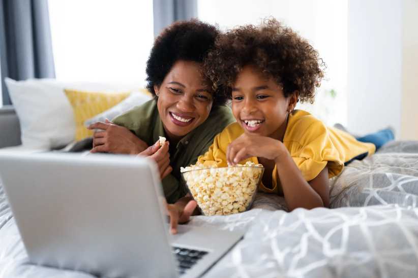 Gorgeous smiling African American single mother enjoying free time with daughter while watching funny videos online on laptop and lying on bed. Black History Month