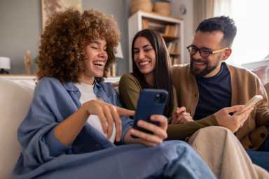 Candid portrait of three diverse young adults in casual sweaters sharing mobile phone content, displaying genuine laughter and joy while lounging on a gray couch in modern living room setting.