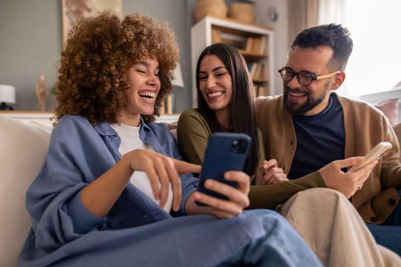 Candid portrait of three diverse young adults in casual sweaters sharing mobile phone content, displaying genuine laughter and joy while lounging on a gray couch in modern living room setting.