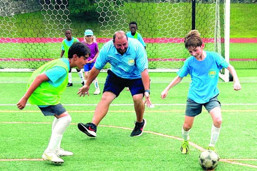Children and a coach play soccer on a green field, smiling and actively engaged near the goal during practice or a camp.
