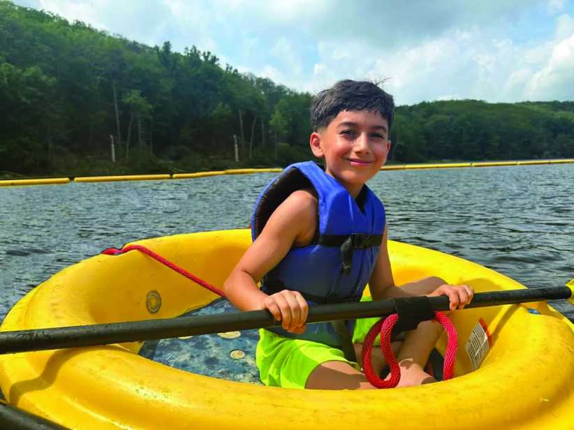 A smiling boy wearing a life vest sits in a yellow boat on a lake, holding a paddle, with forested hills in the background.