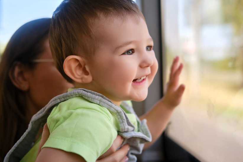 A happy toddler boy looks out the bus window while sitting in the arms of his mother. Child in transport with a parent, close-up. 5 Family Getaways from NYC You Can Do Without a Car