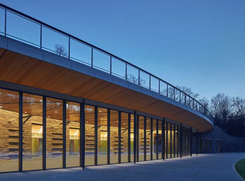 A modern building with large glass windows and a sleek wooden overhang is illuminated at dusk, surrounded by trees and open sky.