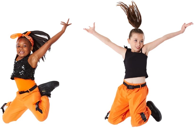 Two girls, wearing orange pants and tops, joyfully leap against a white background.  One girl wears a black sequined top and a headband.
