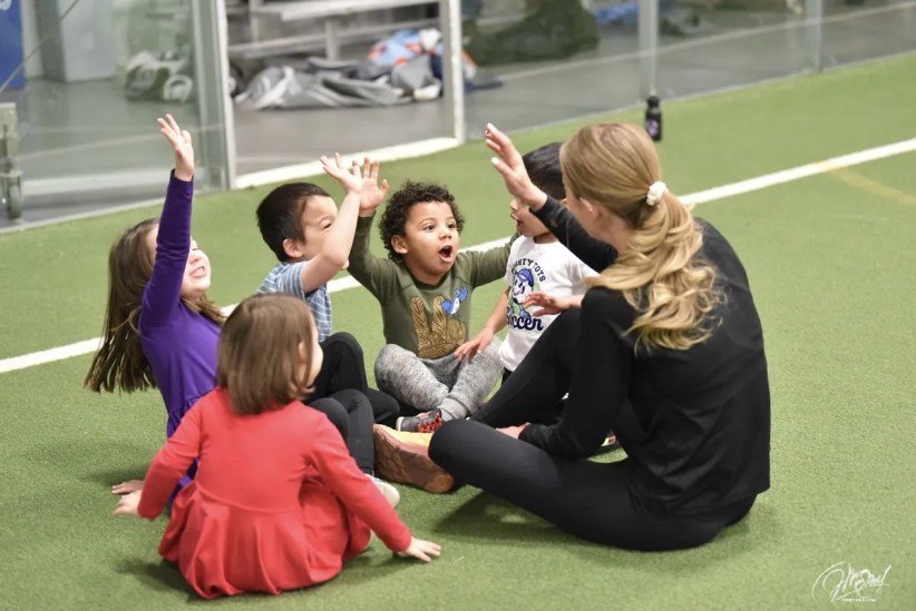 Young children and a woman are sitting on a green indoor playing field. They are playing a game involving raised hands.  One child's shirt says "Mighty Toys Soccer".