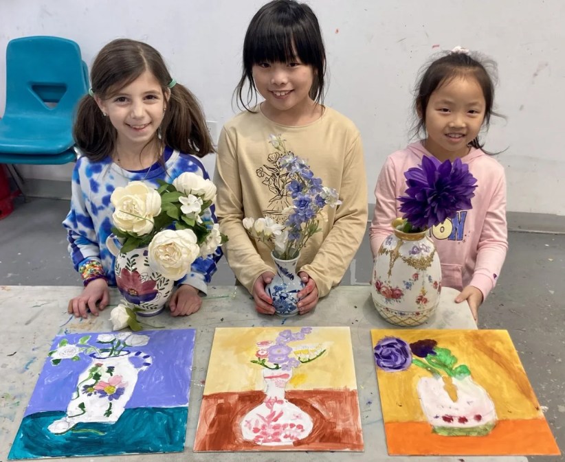 Three girls proudly display floral still-life paintings next to their artistic inspiration: vases of flowers.  The context is an art class setting.
