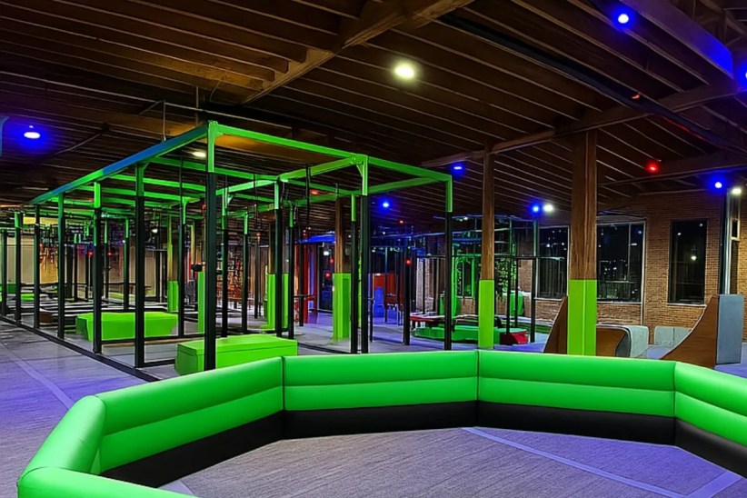 An indoor obstacle course, brightly lit in green and blue, is situated within a large warehouse-like space with exposed wooden beams and brick walls.  A large, padded green seating area is in the foreground.

