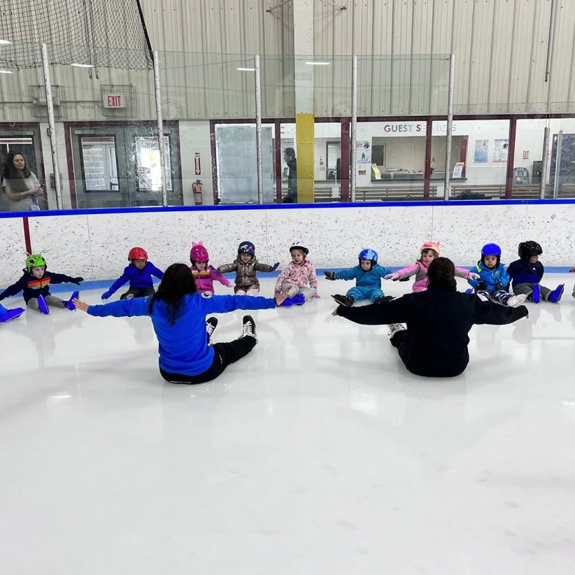 A skating instructor sits on an ice rink, arms outstretched, guiding a circle of toddlers wearing helmets.  They are all seated on the ice.  The background shows an indoor rink and a "GUEST SERVICES" sign.
