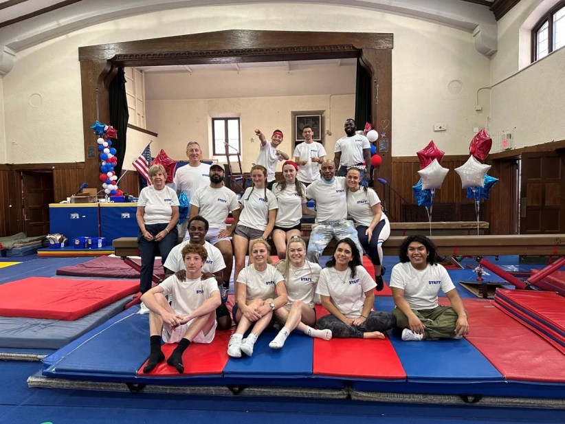 A diverse group of smiling camp staff—men and women of various ages—pose together on stacked gym mats inside a large, wood-paneled multi-purpose room.