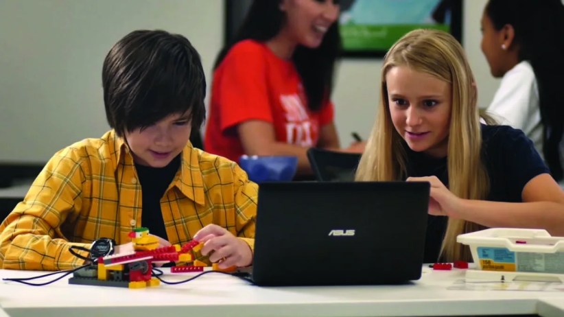 Two children construct a Lego robot at a table using a laptop; other students and a teacher are visible in the background of a classroom.  The laptop's brand is "ASUS".
