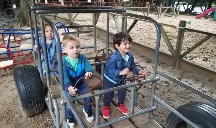 Three young children play together on an outdoor playground structure shaped like a car, smiling and pretending to drive.
