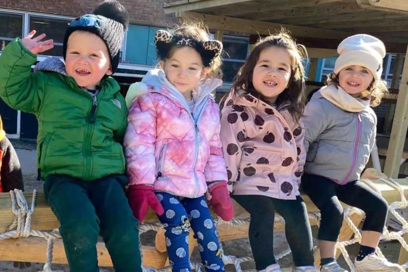 Four young children in colorful winter jackets and hats sit together on a wooden and rope play structure outdoors, smiling happily.