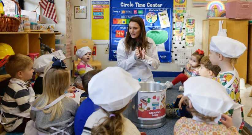 A teacher leads a group of young children wearing chef hats in a colorful classroom, gathered around a large Alphabet Soup bucket for an activity. A young child in a striped swimsuit and large sunhat plays on the sandy beach, using a shovel and Hello Kitty bucket.