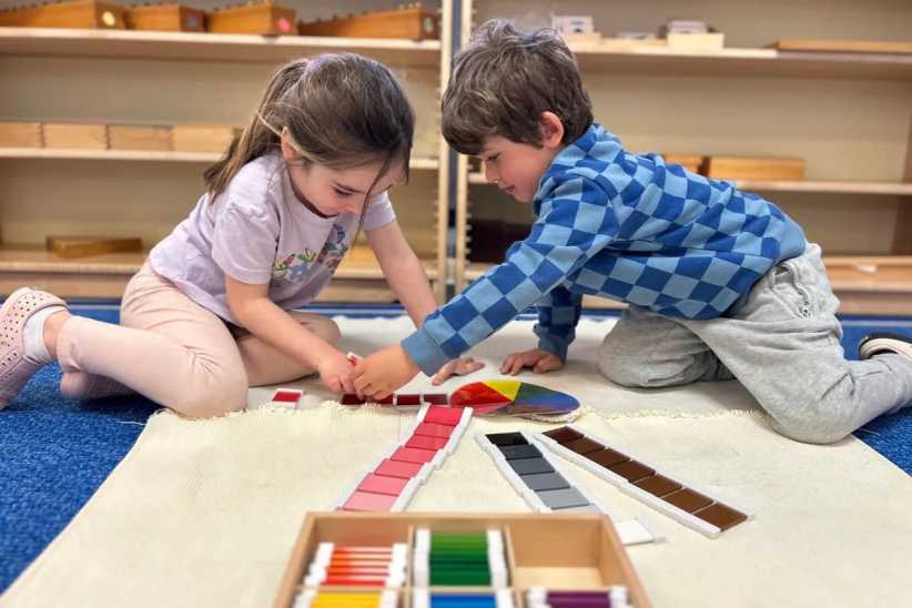Two young children sit on a mat in a classroom, collaborating while arranging colorful tiles and swatches in rows for a learning activity.