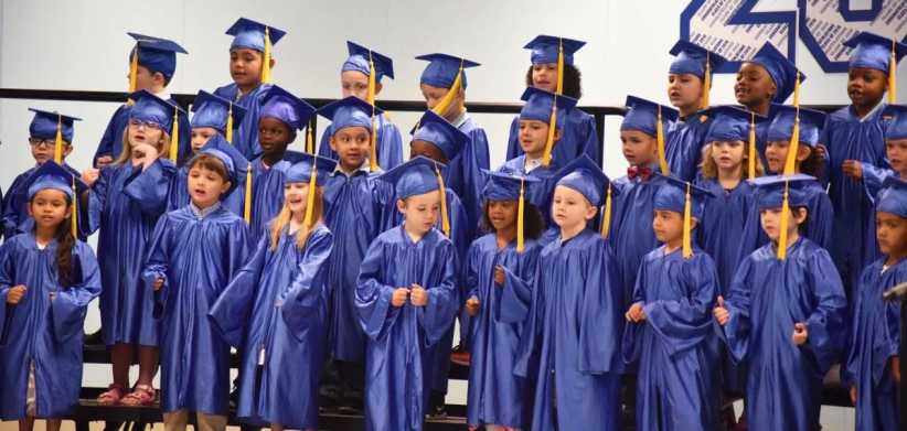 A large group of young children dressed in blue graduation gowns and caps stand together on risers, celebrating their graduation ceremony.