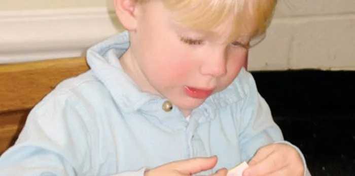 A young child with light hair, wearing a light blue shirt, sits at a table focusing intently on an activity with their hands.A lighthouse stands on a grassy cliff at sunset, overlooking a rocky shoreline and calm ocean under a pastel sky.