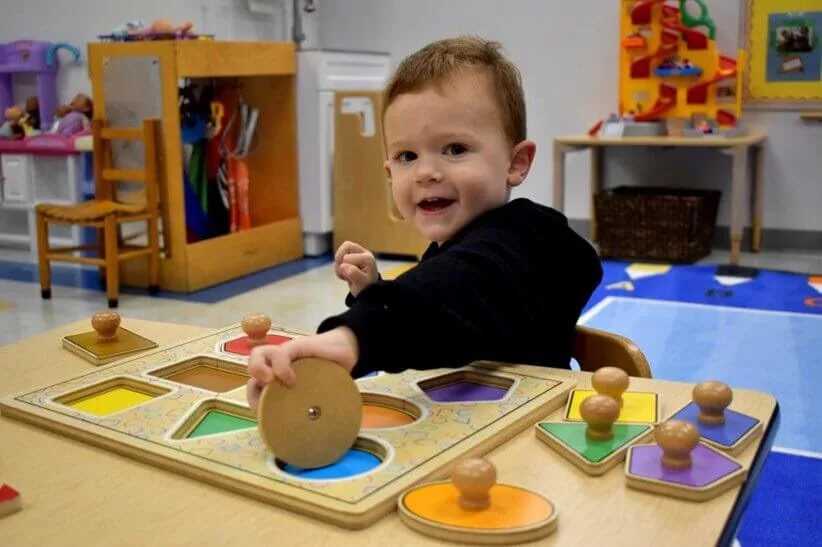 A young child sits at a classroom table, playing with colorful wooden shape puzzles and working on matching shapes and colors.