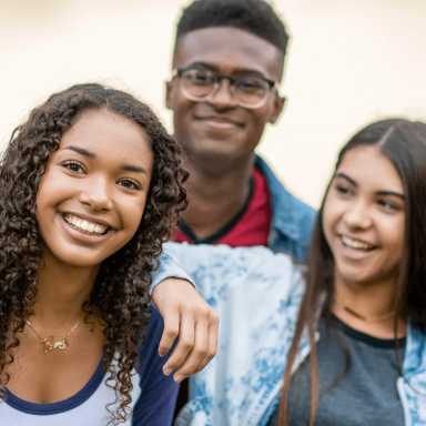 Three teenagers, smiling, pose closely together outdoors. A young woman with curly hair is in the foreground; a young man with glasses and another young woman are behind her. One arm is around the young woman's shoulders.