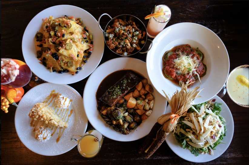 Several dishes are arranged on a dark wooden table; among them are nachos, roasted cauliflower, pasta, a meat dish with potatoes and Brussels sprouts, a dessert, and a salad. Drinks are also present.