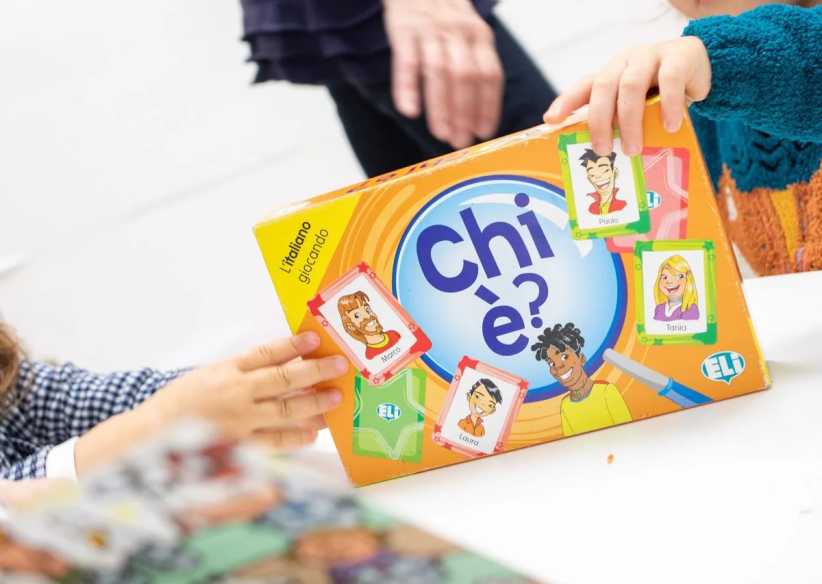Two children holding a colorful Italian board game box titled 'Chi è?' with cartoon character cards attached on the front.