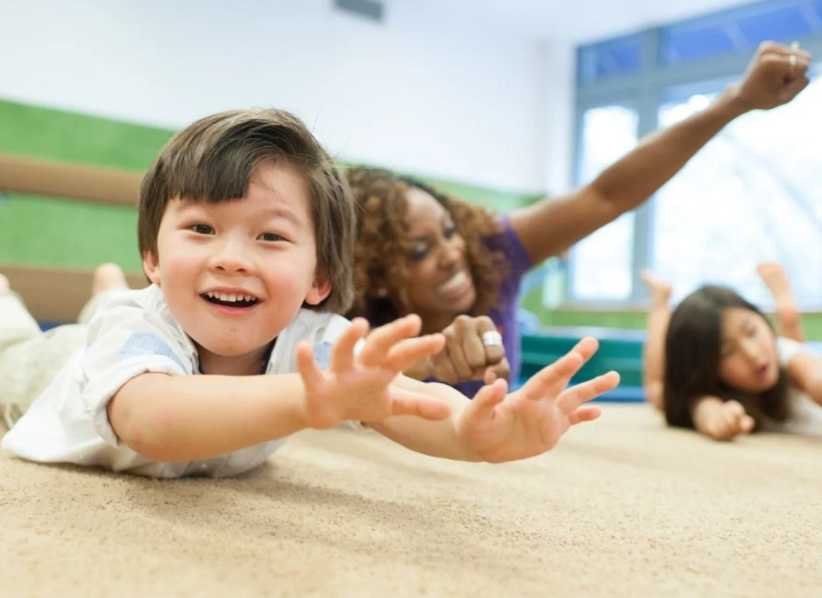 Smiling child lying on the floor reaching forward playfully, with a teacher and other kids in the background.