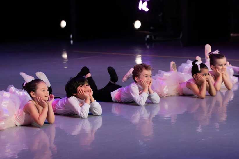 Young children in ballet costumes lying on stage with hands under chins, smiling during a dance performance.